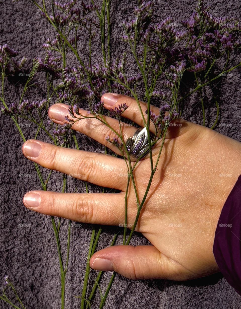 Female hand on a grey coloured cloth with a flower-shaped ring with purple stones and purple flowers between the fingers.