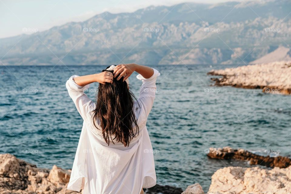 Back view of a young woman in a white shirt standing on the rocky coast of the sea