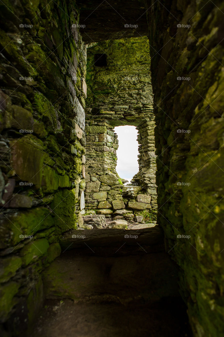 Passageway with square and rectangle stone walls and rectangle window at the end of the tunnel. Shapes squares and rectangles. Green moss on walls.