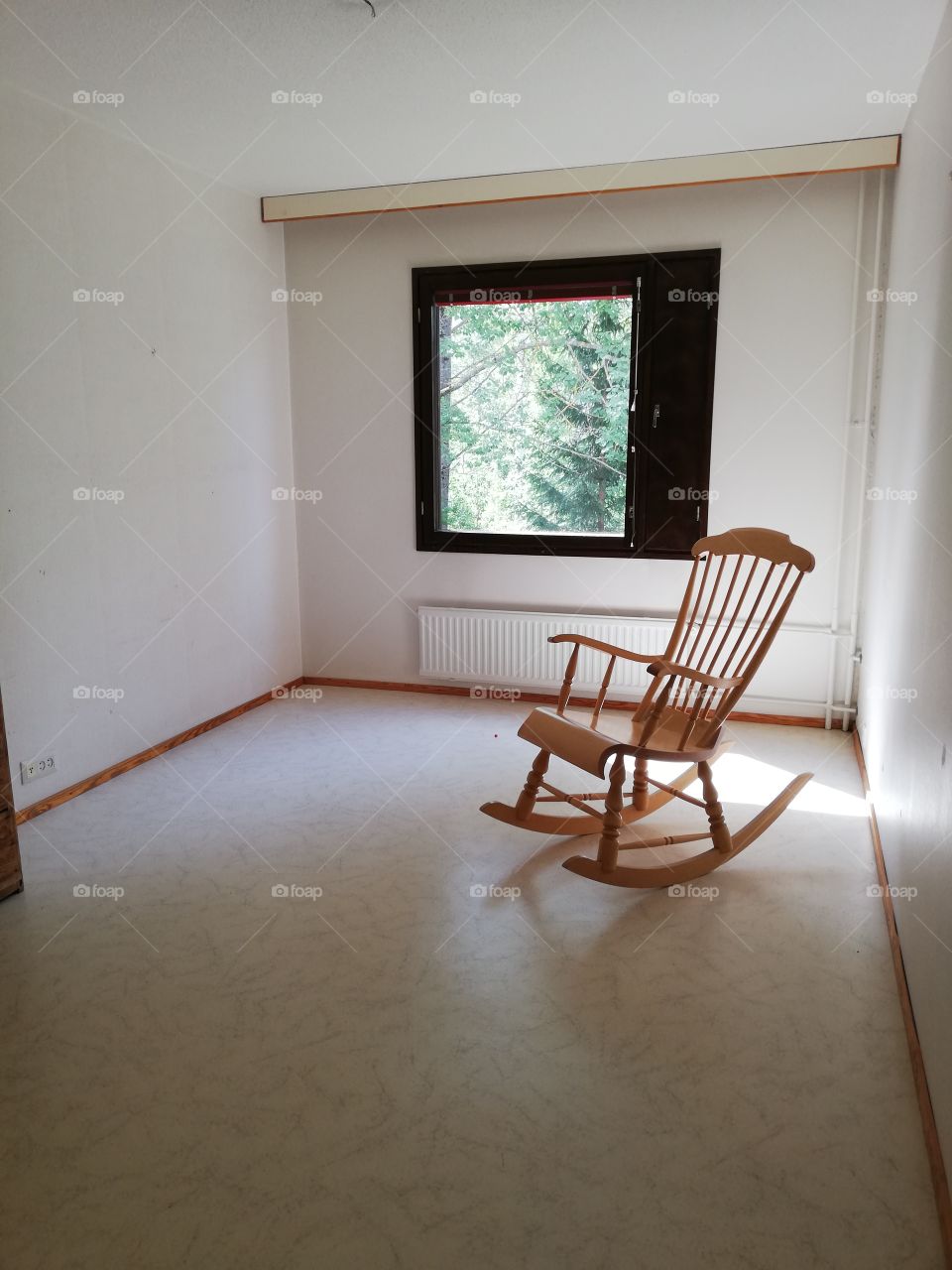 A beige rocking chair made of varnished wood is standing in an empty room. The walls, a ceiling and a heater are white, patterned plastic mat. Light colors all over the room, except brown fortochka and window frames.
