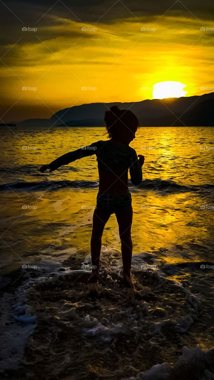 Action photography: Image of child on the beach jumping on a wave on his back.  In the background, sunset and mountains.
Fotografia de ação: Imagem de criança na praia pulando onda de costas.  Ao fundo, por do sol e montanhas.