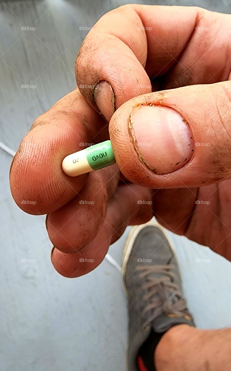 closeup of a rough male hand holding a prescription pill between thumb and index finger . the fingers are stained and dirty. the finger nails are chewed