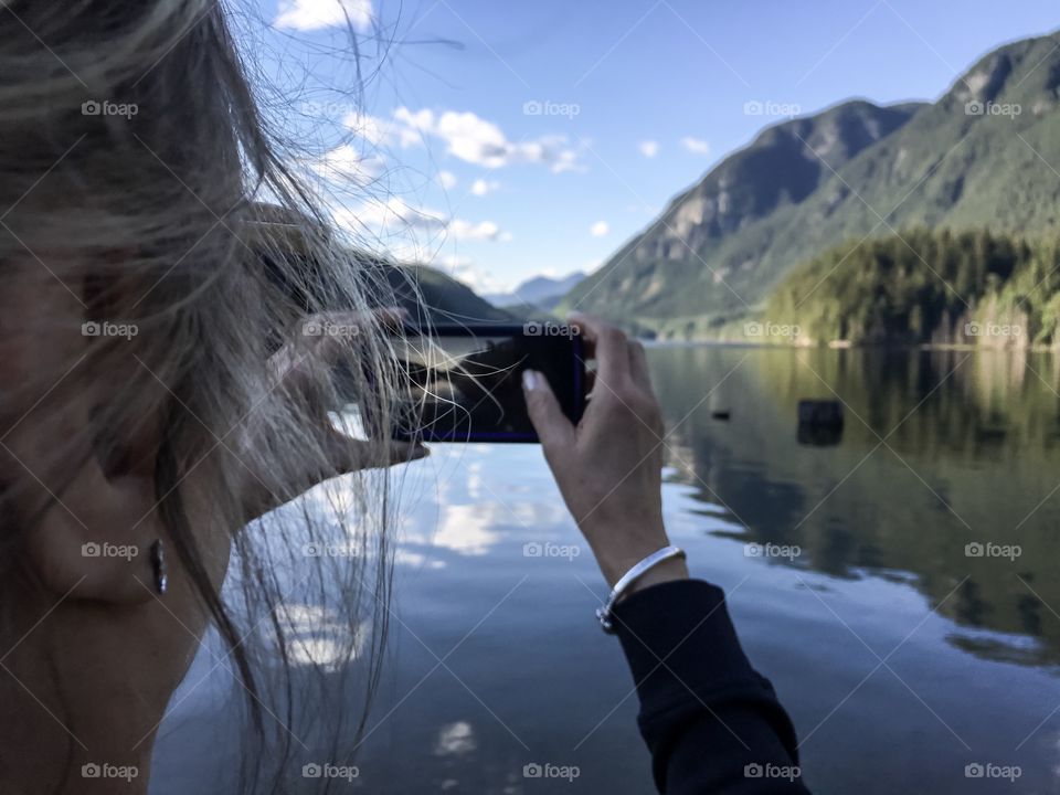 Blonde woman female photographer capturing image of mountain lake and reflections late day, rear view head shot