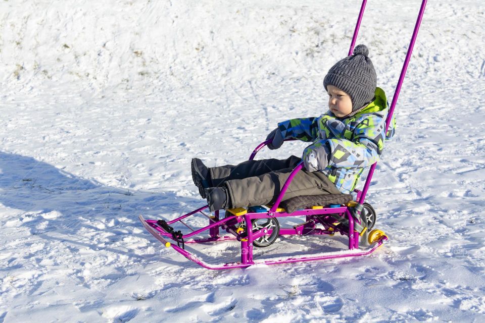 A child in winter in winter jackets, pants, a hat and boots on white snow on the street and in the park in nature plays winter fun and sleds.