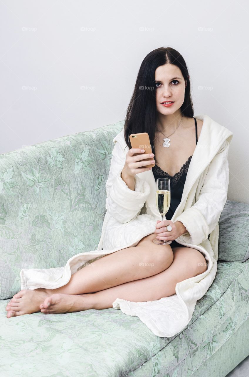 Beautiful luxury brunette girl sit in white robe and black negligee on green sofa with iphone 