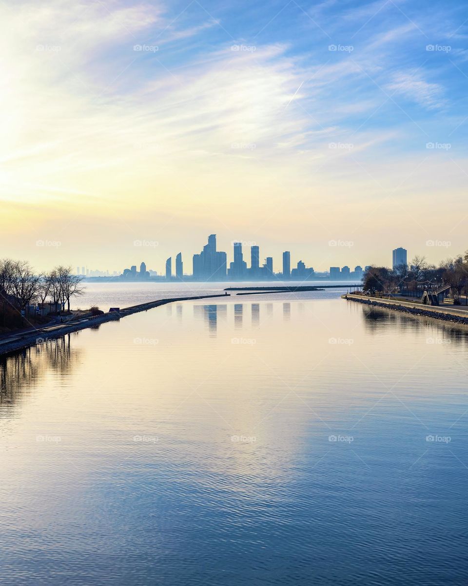 A city skyline reflected in a calm blue lake, with sunset glow lighting up the clouds.