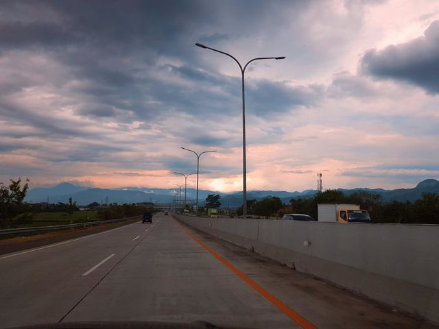 Driving  on the Seroja toll road, Bandung city, on the edge toll road can see rice fields with green leaf tress, lighting poles on road medians, mountains and cloudy skies, yellowish red to gray in color.