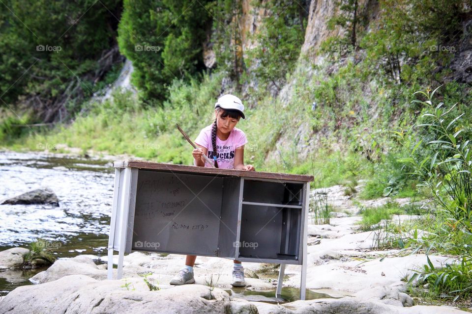 Cute little girl is writing at the desk on a river bank