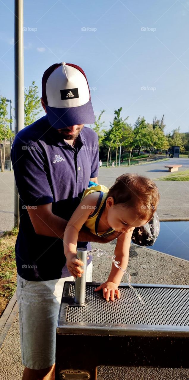 The father is holding the baby and helping him to play with the spring water