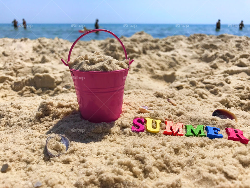 View from a sandy beach by the sea with people on the sand pink bucket with sand and inscription summer