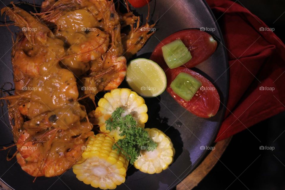 Asian food balado fried prawns with corn chunks, lemon slices and tomatoes served on black plate with red cloth bottom and wooden coaster. Asian style street seafood.