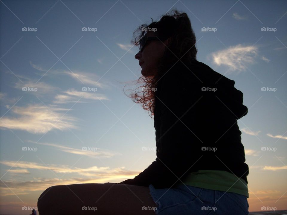 Silhouette of woman thinking sitting against bright blue sky with soft clouds and sunset light at bottom looking at horizon