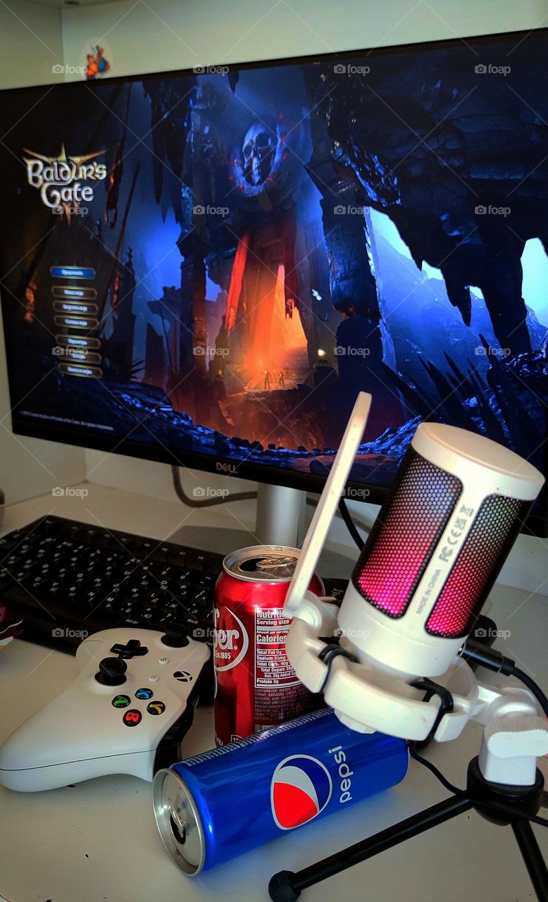 Computer game "Baldur's Gate 3" on a computer monitor. On the table: keyboard, game joystick, Pepsi can, Dr Pepper can and microphone