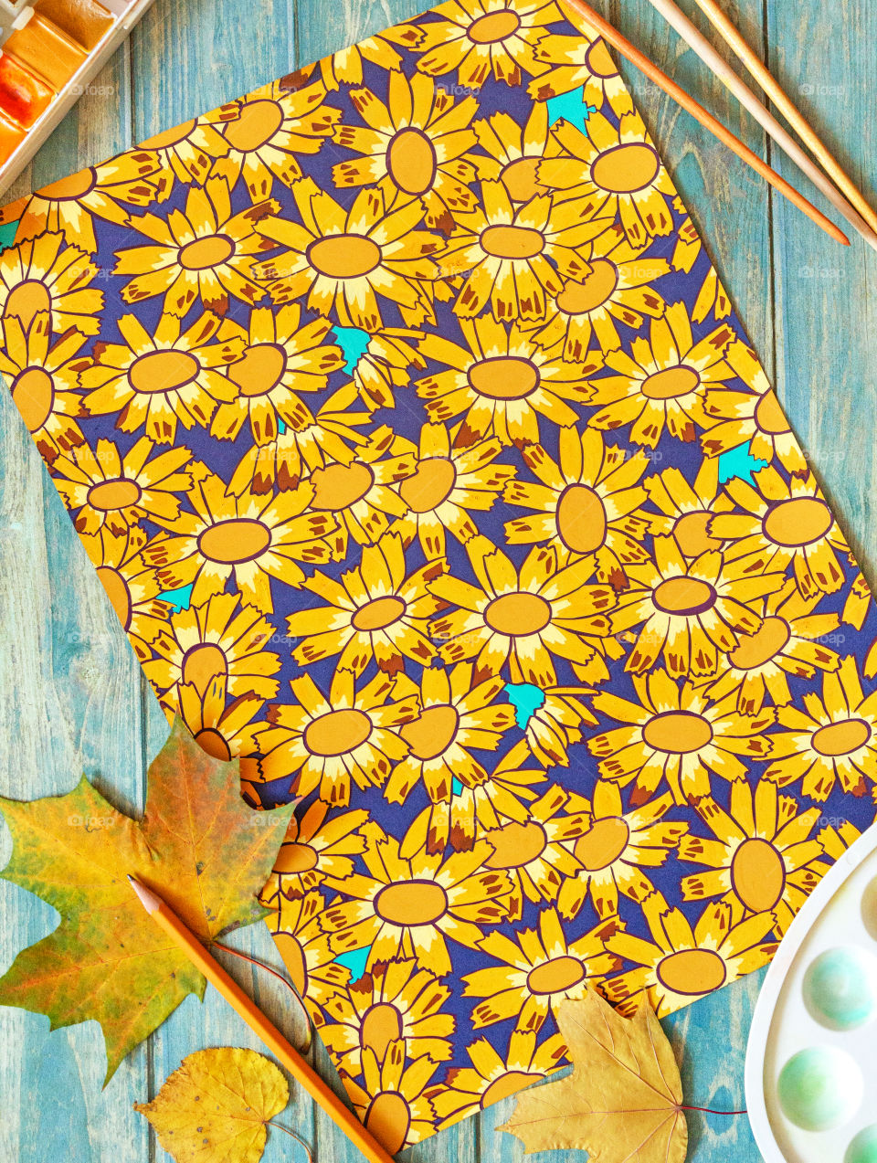 Creative work, paint drawing.  A table with a decorative repeating pattern with daisies, next to which are brushes, watercolor and gouache paints, a palette and dry autumn colored leaves.