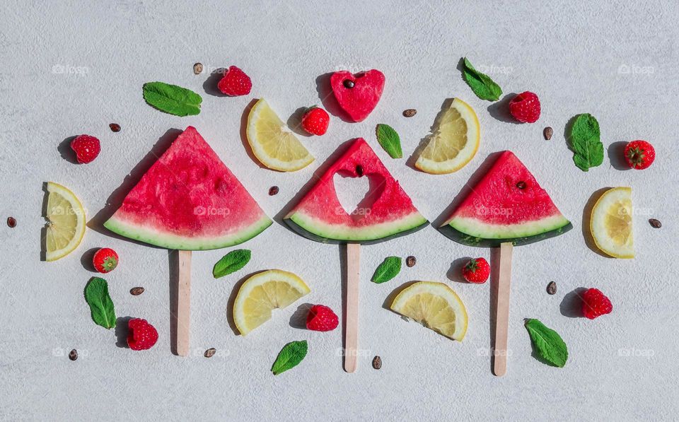 Three slices of watermelon with mint leaves, lemon slices and raspberries on a light gray cement background, flat lay close-up. Concept summer treat,summer food,watermelon mood,healthy lifestyle.