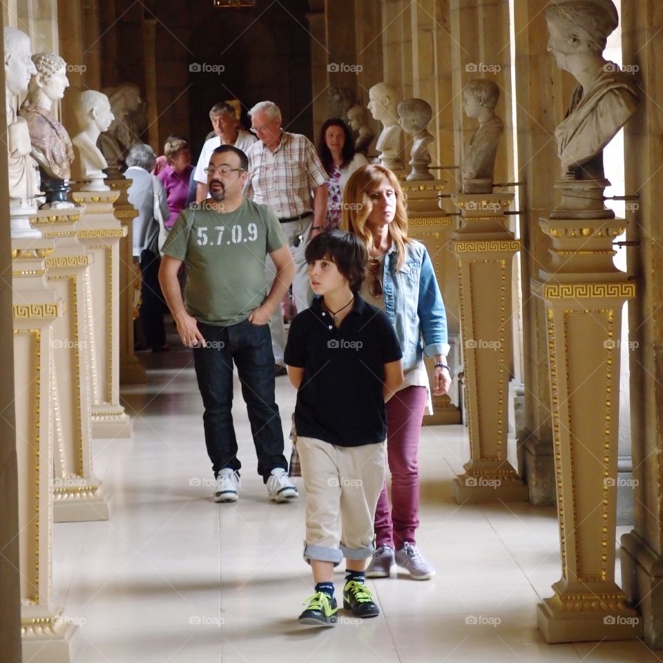 A boy and his family walk through a corridor in a castle exploring the multiple busts perched on pillars against the walls. 