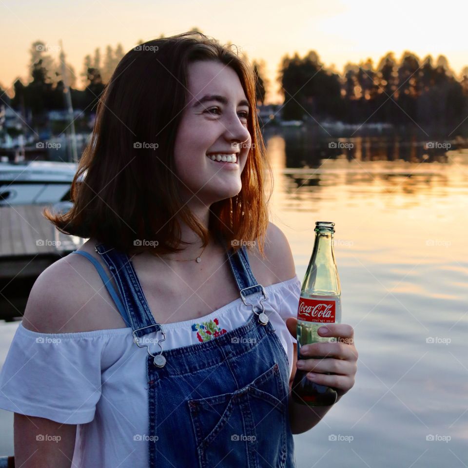 Happy young woman drinks a Coke product while enjoying the sunset on a lake