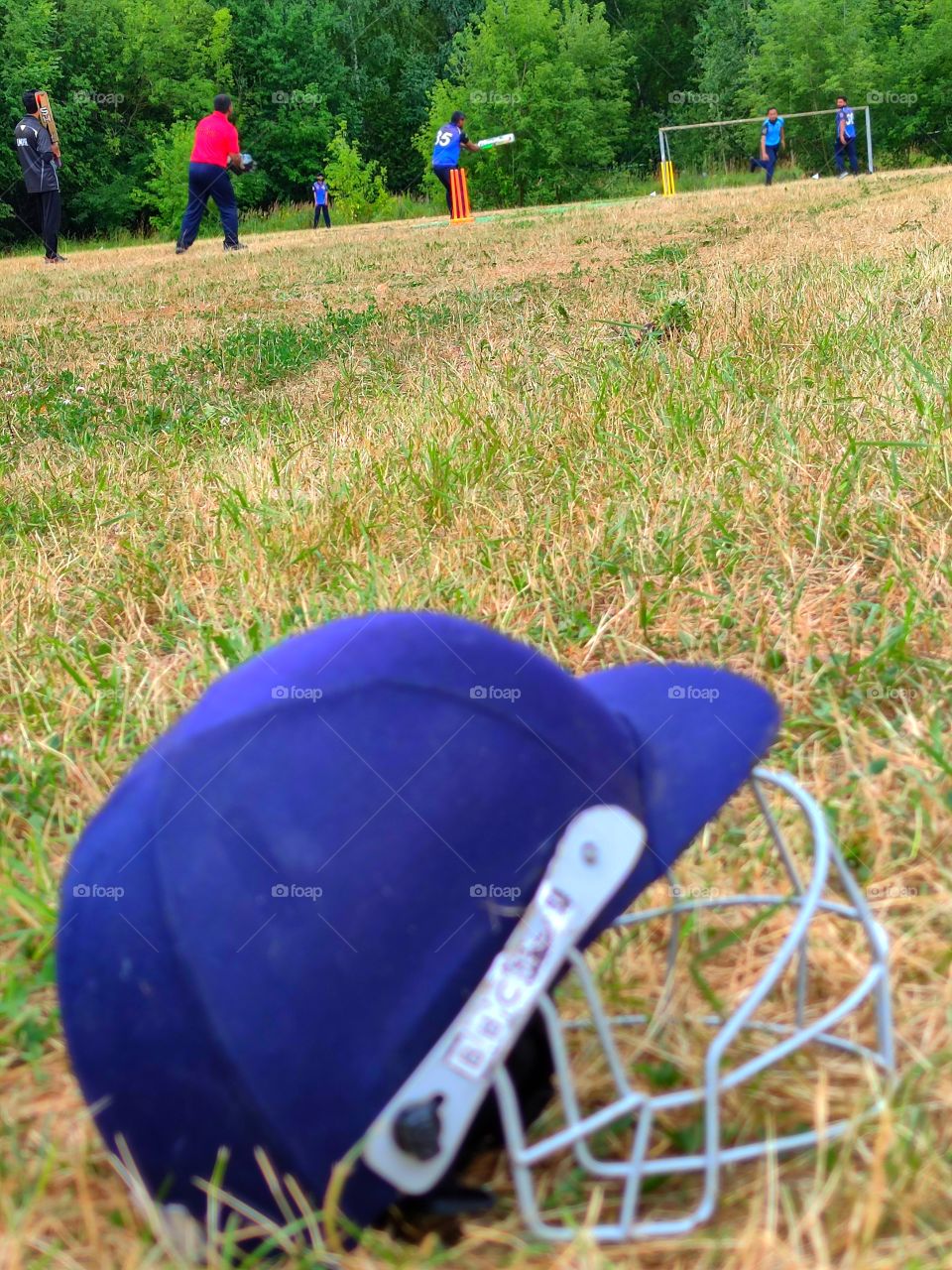 cricket game.  one player in blue is standing up to hit the ball.  the second player threw the ball.  in the foreground of the photograph lies on the field a protective helmet for cricket
