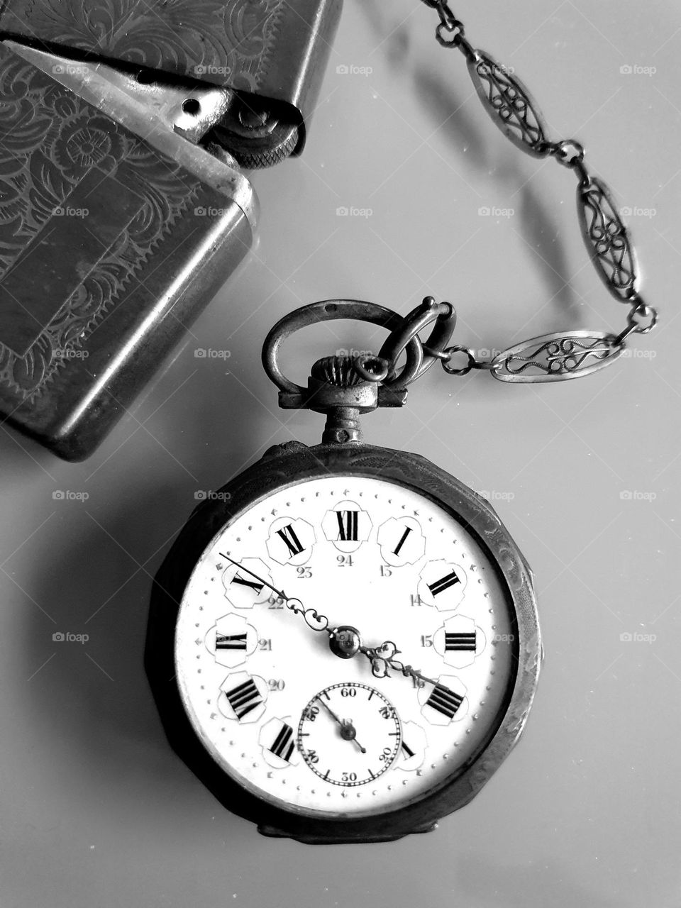 Black and white fatlay composition on a grey glass surface of a vintage pocket watch and a zippo lighter