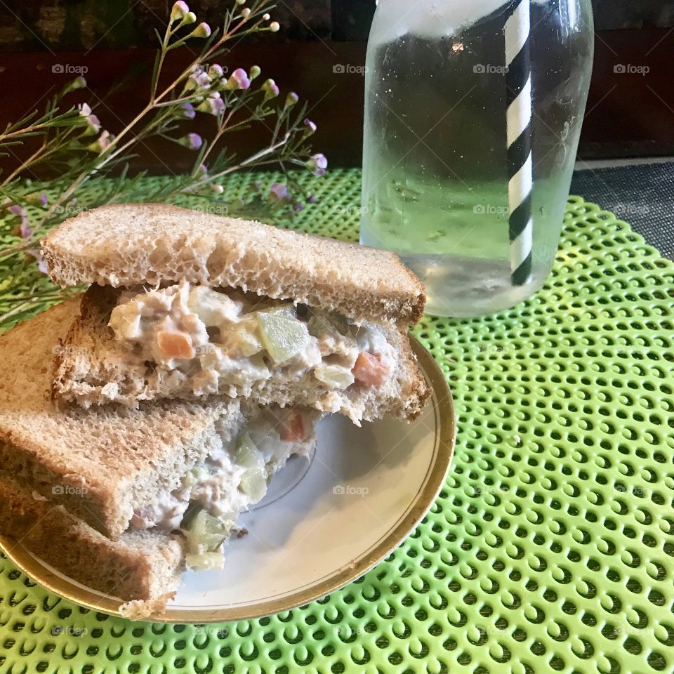 A very healthy tunafish salad sandwich On white bread with celery, pickles and red bell peppers with a jug of ice water with a black and white straw, lavender flowers on a lime green placemat.