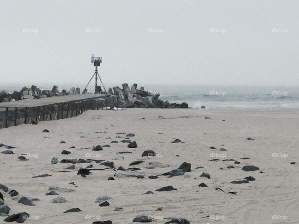 A photo of a beach in Point Pleasant Beach, NJ near the Manasquan Inlet. Rocks in the foreground make the beach resemble the moon, as does the gray sky. On the left is a pier people walk out on to fish or to just get a closer view of the ocean.