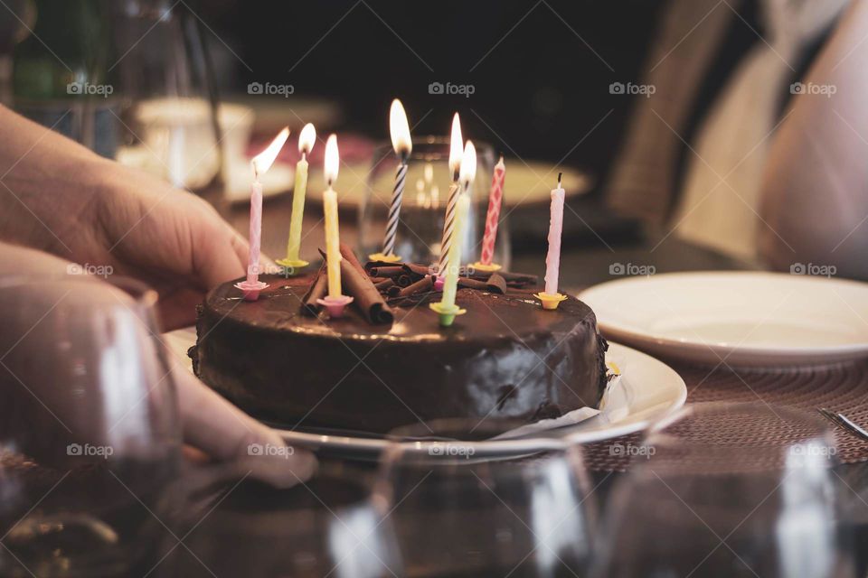 A portrait of someone putting a chocolate birthday cake on a table with some burning candles on it. some of them went out when carrying it. the lit candles need to be blown out at once in order to get a birthday wish.