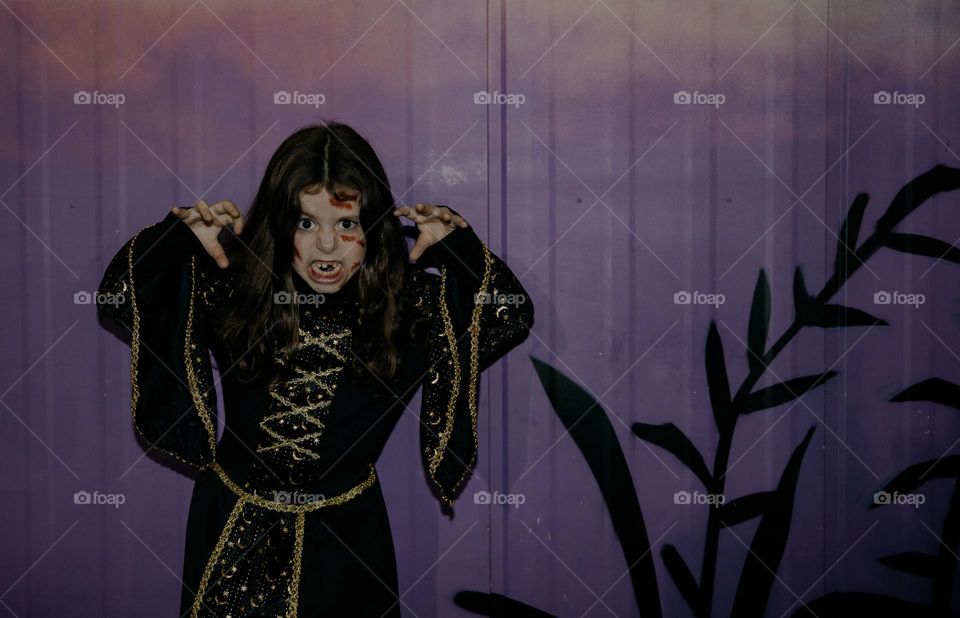 Portrait of a beautiful little Caucasian brunette girl in a witch dress with Halloween makeup stands against the background of a purple wall with raised hands and a frightening emotion on her face late in the evening on the street, close-up side view