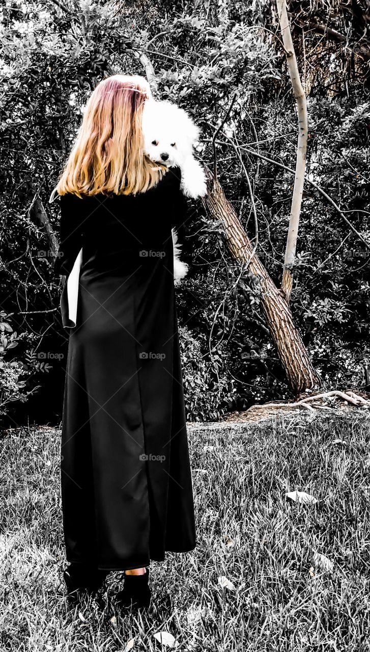 In a contrast of color, a white puppy peers over the shoulder of a blond girl wearing a long black dress in a wood.