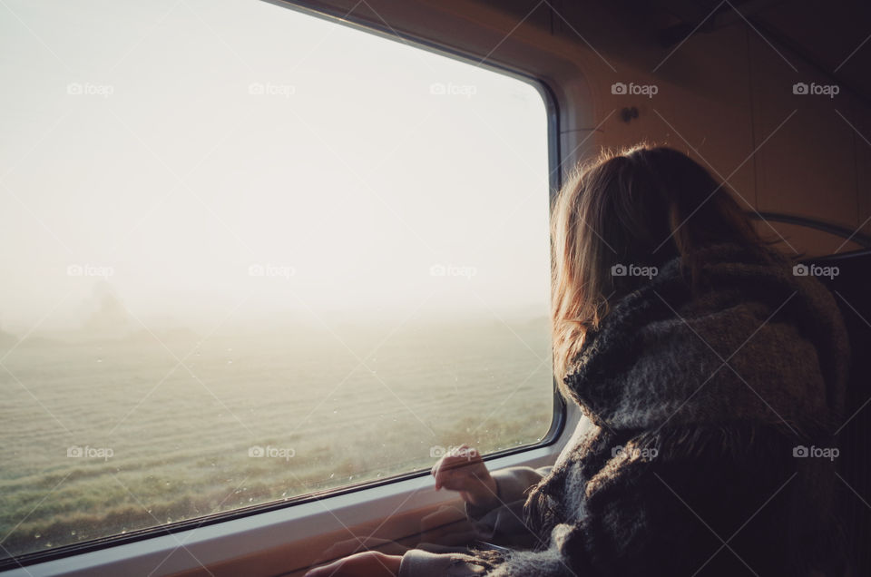 Girl looking out of the train's window into the foggy sunrise over the field.