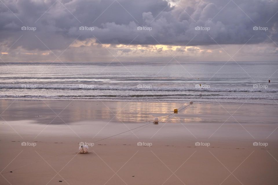Moments before sunrise on a golden beach, between the gentle clouds, a soft glow emerges. The sand takes on salmon hues, and the buoys mark an area for boats