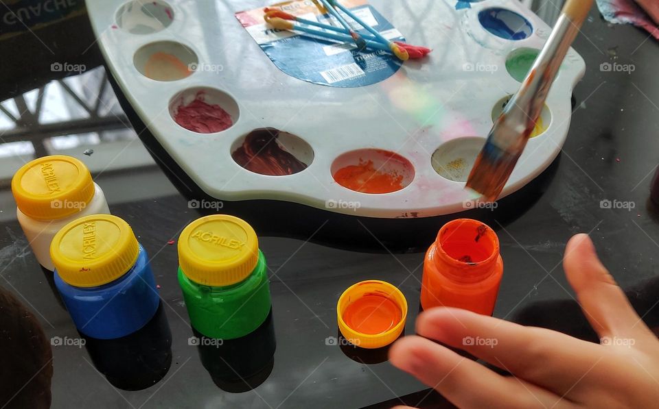 A child's hand picks up red paint with a brush.  Nearby on a black mirrored table lies a palette and cans of green and blue paint.