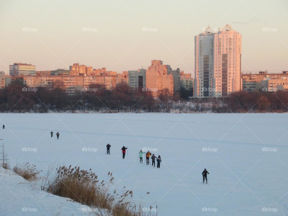 A group of skiers is walking along a frozen river, on snow and ice, winter, cold, frost, evening