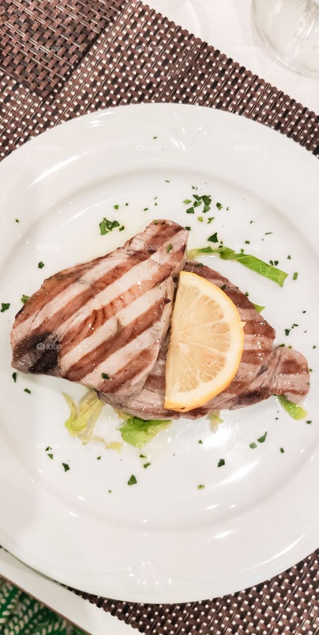 Grilled tuna with a slice of lemon in a white plate on a table in a restaurant in italy, flat lay close-up. Sea food concept.