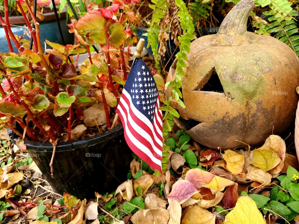 Small U.S.A flag in planter next to outdoor garden pumpkin pottery surrounded by fall leaves