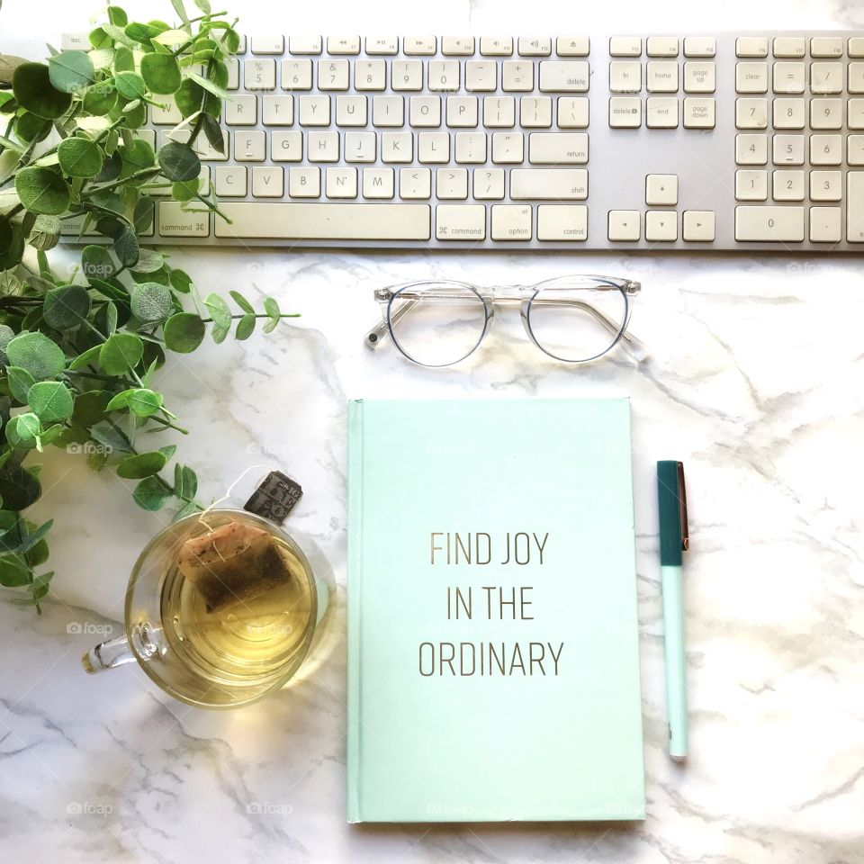Find the joy in the ordinary in your daily lives! Green book on a marble table with clear glass with tea and a white keyboard!