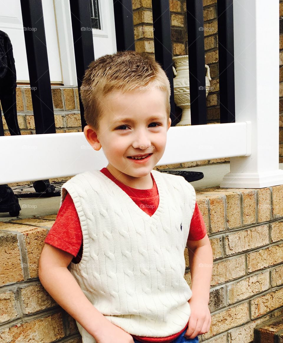 Cutest face ever! Smiling little boy outside in front of his home in adorable sweater vest and red t-shirt. 