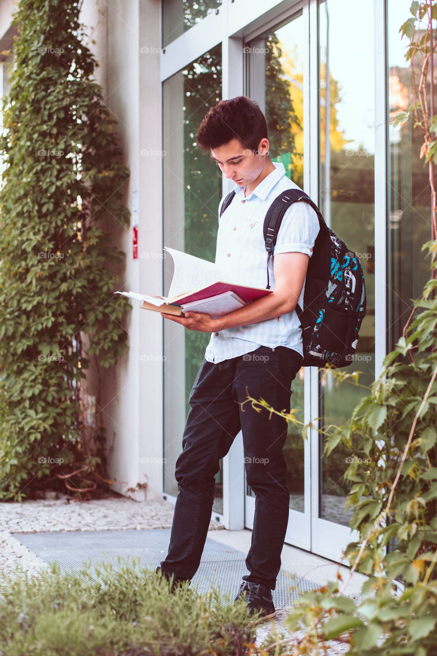 Student holding a notebook and carrying a backpack standing at the front of university building. Young boy wearing blue shirt and dark jeans