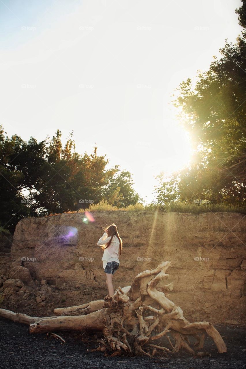 A girl on the shore in the rays of the setting sun stands on a snag