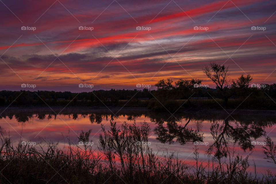 Just before dawn the morning sky above this pond is glowing with deep red pink orange and blue colors which are reflecting in the waters surface