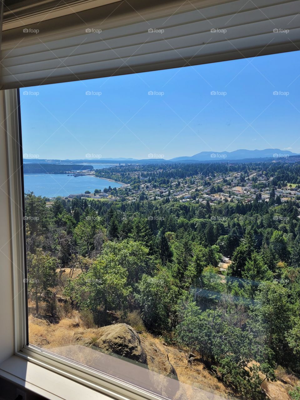 Looking through uphill home window to clear blue skies, lush trees, rocks, islands, mountains, and pacific ocean views of Nanaimo on Vancouver Island, Canada