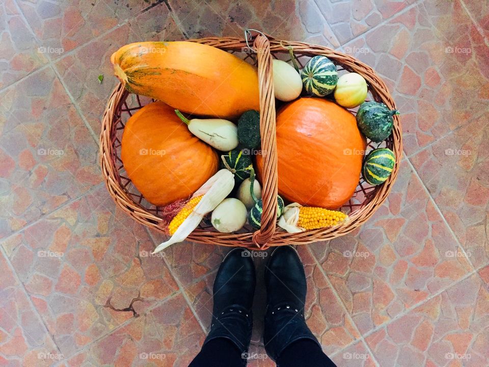 Looking down at black boots near a wooden basket with different pumpkins and dry corn