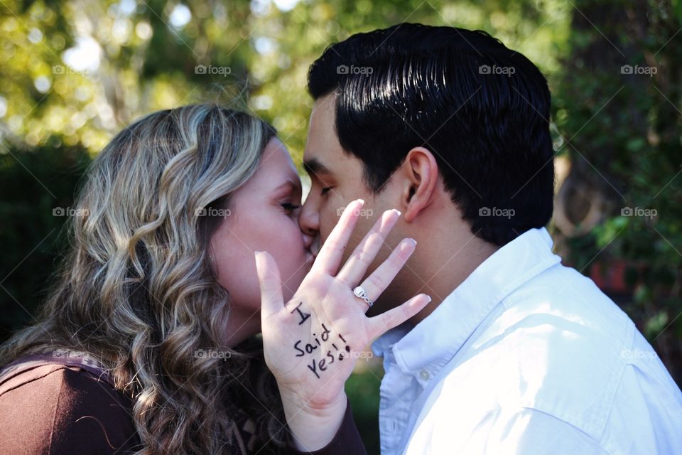 An engagement photo of the couple kissing with the bride showing off her ring and the expression, "I said Yes!" Written on her hand! 