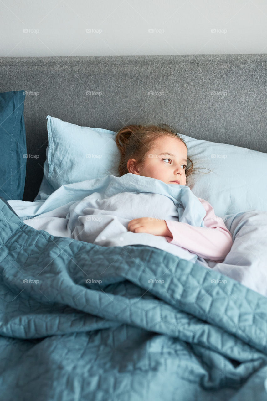 Ill little girl lying in bed covered with duvet. Sick kid recovering, resting in bed, looking at window