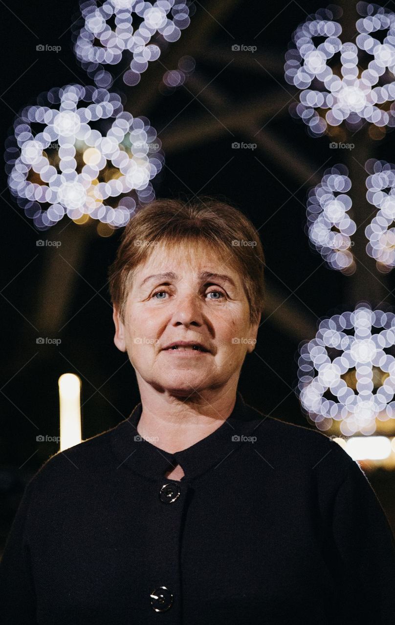 Portrait of one elderly beautiful Caucasian woman walking at night against the backdrop of a blurry glowing atomium in Brussels and looking at the camera, close-up side view