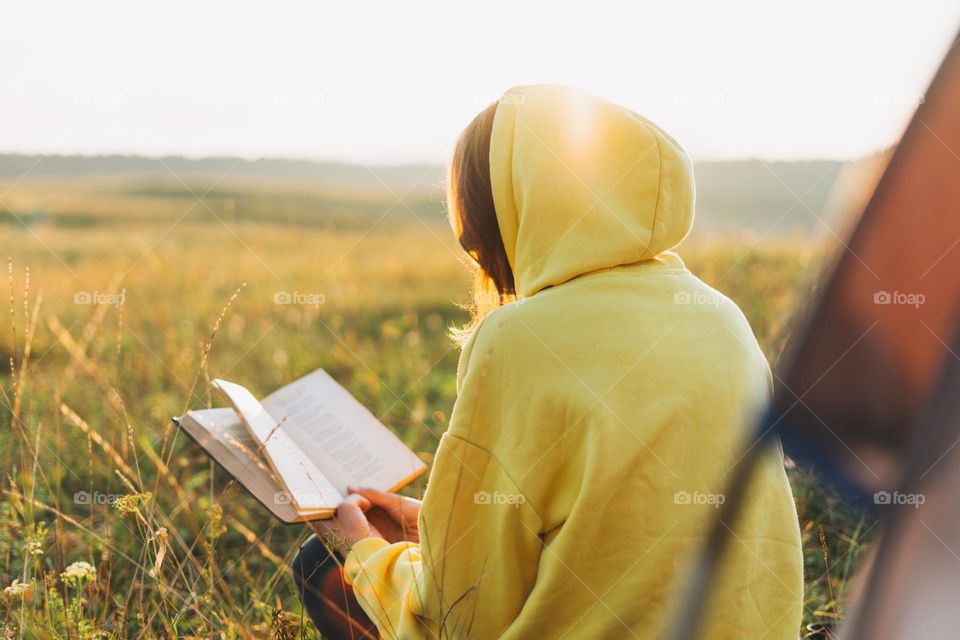 Young woman traveler in yellow hoodie reading book against tent. Local tourism, weekend trip