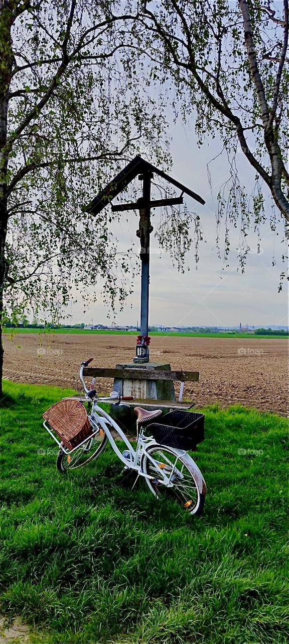 This country road in rural “Bavaria”, Germany forks off in several directions from this rest stop with a bench and a crucifix above it. 2024. Hypnotic Productions