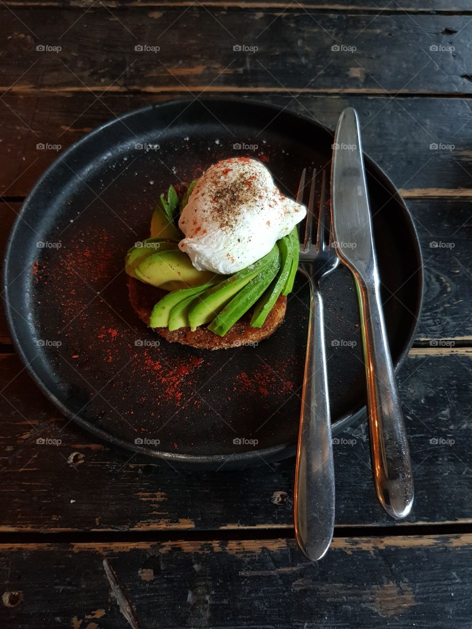 Delicious & beautiful avocado toast with poached egg on a black plate; fork and knife are lying next to it. All on a wooden table.