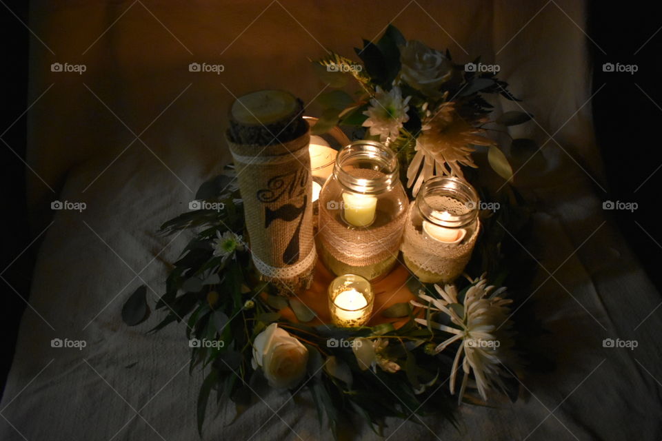 A wedding center piece comprised of willow and eucalyptus greens, white flowers and warm candle light are as romantic as it gets. The table number and candle jars adorned with burlap and lace add a delicate touch. Lit with candle and ambient light 
