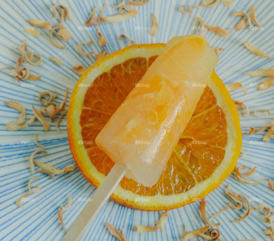 A frozen ice lolly on a slice of orange, surrounded by dried orange peel on a blue patterned plate