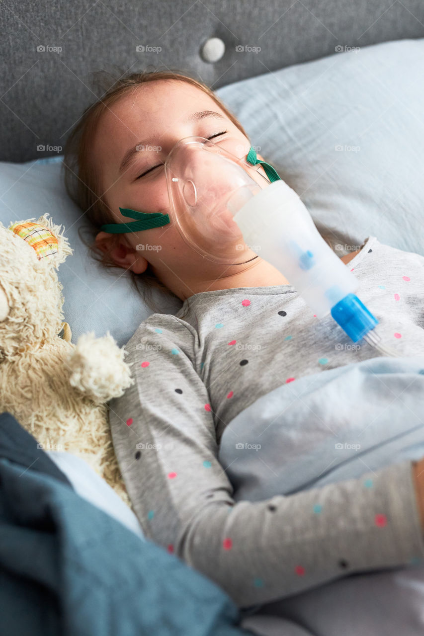 Sick little girl having medical inhalation treatment with nebuliser. Child with breathing mask on her face lying in bed